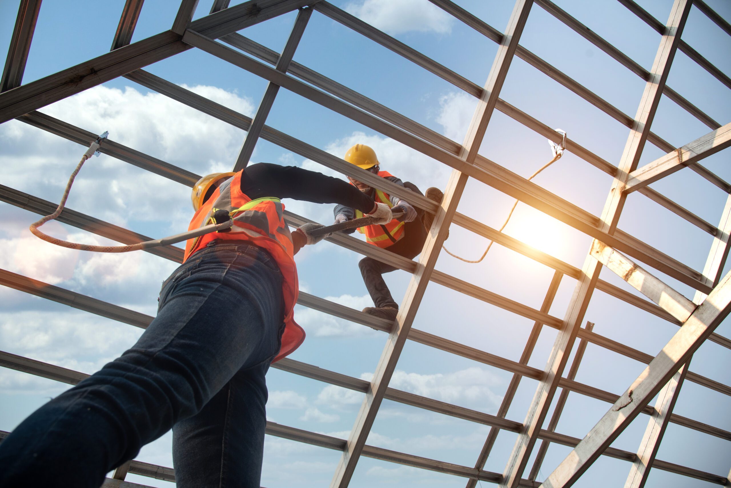 Construction,Workers,Install,New,Roofs,,Roofing,Tools,And,Fall,Protection Two construction workers wearing safety gear are working on a metal structure with a blue sky and some clouds in the background.